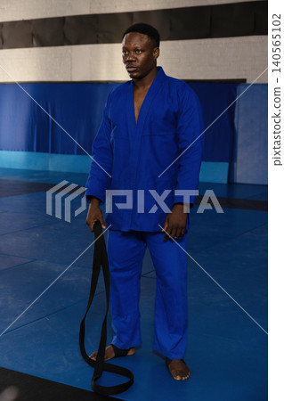 African american man in blue gi standing barefoot on mat holding belt in martial-arts hall African american man in blue gi standing barefoot on mat holding belt in martial-arts hall 140565102