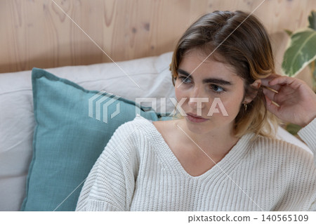 Woman wearing knit sweater sitting on couch tucking hair in living room with teal cushion, plant Woman wearing knit sweater sitting on couch tucking hair in living room with teal cushion, plant 140565109