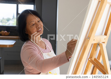 Senior asian woman painting on canvas at wooden easel at home with paintbrush, splattered apron Senior asian woman painting on canvas at wooden easel at home with paintbrush, splattered apron 140565124