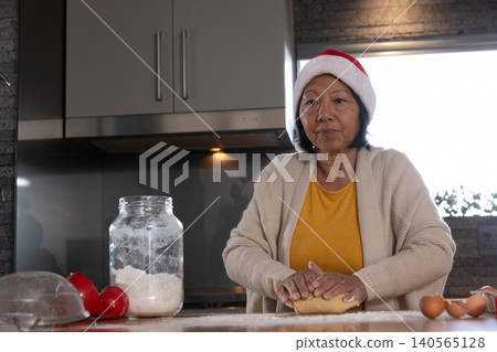Senior Asian woman kneading dough on wooden countertop in kitchen with flour jar and Santa hat Senior Asian woman kneading dough on wooden countertop in kitchen with flour jar and Santa hat 140565128