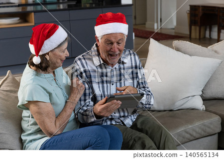 Senior couple sitting on sofa near kitchen cabinets pointing at tablet wearing Santa hats Senior couple sitting on sofa near kitchen cabinets pointing at tablet wearing Santa hats 140565134