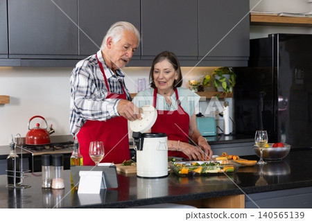 Senior couple wearing red aprons operating food processor and slicing vegetables at kitchen island 140565139