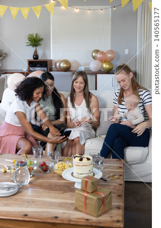 Diverse female friends chatting and holding infant on sofa at home with cake, pastel balloons Diverse female friends chatting and holding infant on sofa at home with cake, pastel balloons 140565177