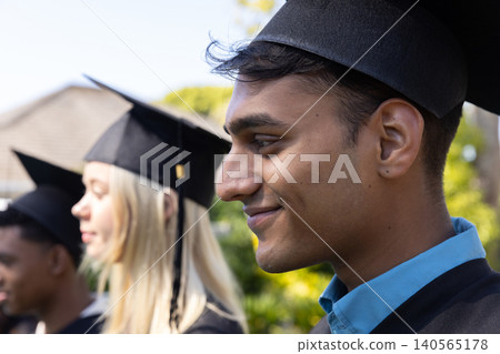 Diverse graduates standing in line on campus wearing mortarboards, gowns against foliage Diverse graduates standing in line on campus wearing mortarboards, gowns against foliage 140565178