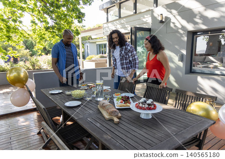 Diverse friends gathering around wooden dining table on shaded backyard deck with cake, balloons Diverse friends gathering around wooden dining table on shaded backyard deck with cake, balloons 140565180