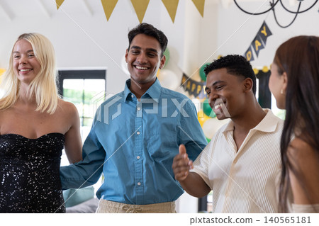 Diverse friends standing in living room, linking arms under happy birthday banner, balloons 140565181