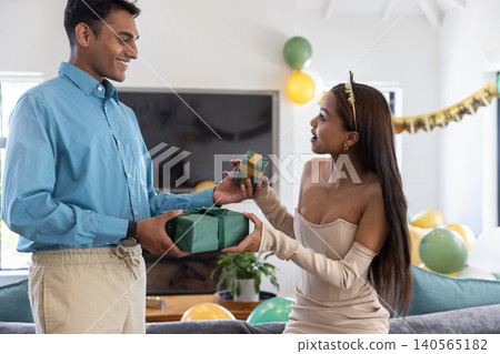 Diverse couple exchanging wrapped gifts near sofa in living room with balloons, banner Diverse couple exchanging wrapped gifts near sofa in living room with balloons, banner 140565182