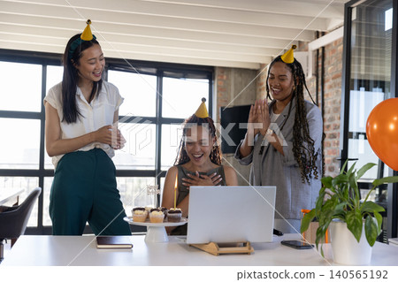Diverse female coworkers celebrating birthday at desk wearing party hats with laptop and cupcakes Diverse female coworkers celebrating birthday at desk wearing party hats with laptop and cupcakes 140565192