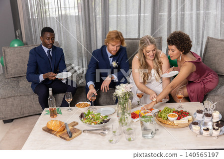 Diverse wedding party in formal attire sharing charcuterie board, pouring rosé at coffee table 140565197