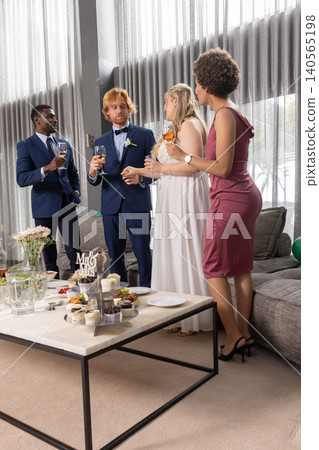 Diverse wedding party in formal wear standing around coffee table in lounge Diverse wedding party in formal wear standing around coffee table in lounge 140565198