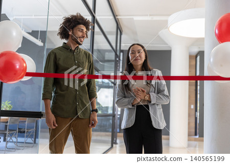 Diverse coworkers in business attire standing behind ribbon in reception area with balloons Diverse coworkers in business attire standing behind ribbon in reception area with balloons 140565199