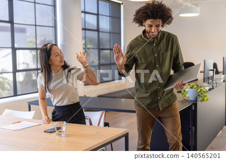 Diverse coworkers exchanging high-five beside table in open-plan office with laptop, smartphone Diverse coworkers exchanging high-five beside table in open-plan office with laptop, smartphone 140565201