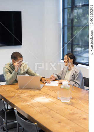 Diverse coworkers sitting in office reviewing documents on laptop, writing, pen near pitcher Diverse coworkers sitting in office reviewing documents on laptop, writing, pen near pitcher 140565202