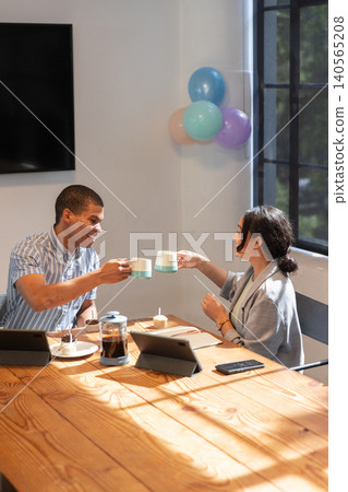 African american man and asian woman clinking mugs in conference room, tablets and coffee press African american man and asian woman clinking mugs in conference room, tablets and coffee press 140565208