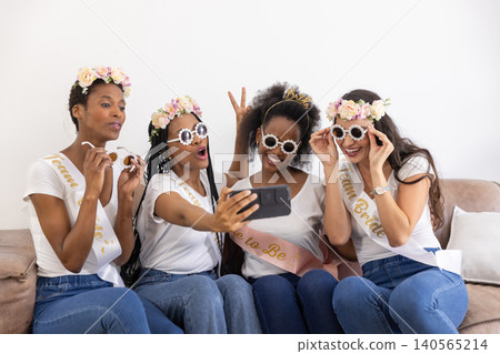Diverse female friends posing for selfie on sofa wearing sashes sunglasses while holding smartphone Diverse female friends posing for selfie on sofa wearing sashes sunglasses while holding smartphone 140565214