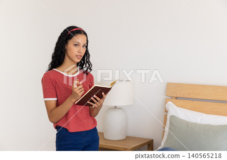 Female teenager standing near lamp in bedroom holding notebook, pen, studying notes, copy space Female teenager standing near lamp in bedroom holding notebook, pen, studying notes, copy space 140565218