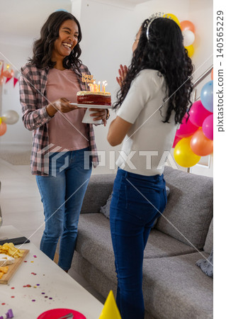 Diverse mom and teenage daughter holding birthday cake with lit candles at home near balloons Diverse mom and teenage daughter holding birthday cake with lit candles at home near balloons 140565229