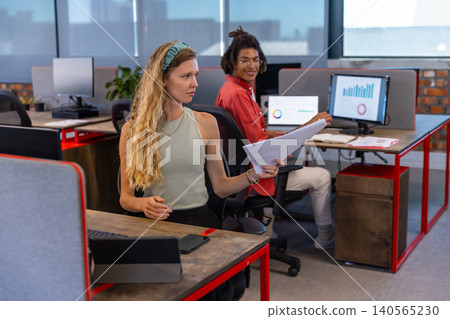 Diverse coworkers reviewing reports at red-framed desks in modern office with monitors, copy space Diverse coworkers reviewing reports at red-framed desks in modern office with monitors, copy space 140565230