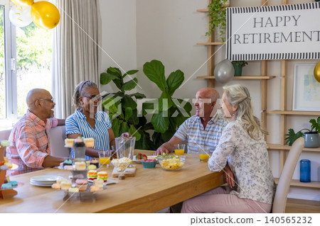 Diverse senior friends celebrating retirement at home dining table with cupcake stand, banner Diverse senior friends celebrating retirement at home dining table with cupcake stand, banner 140565232