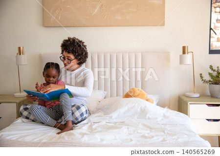 African american mother with daughter reading kids book on tufted bed with teddy bear, copy space African american mother with daughter reading kids book on tufted bed with teddy bear, copy space 140565269