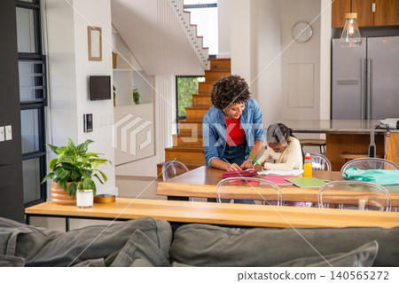 Mother and african american daughter working on homework at dining table with notebooks, pencils Mother and african american daughter working on homework at dining table with notebooks, pencils 140565272
