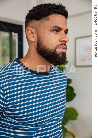 Adult man standing looking right in home wearing striped tee by black-framed window with plant Adult man standing looking right in home wearing striped tee by black-framed window with plant 140565499
