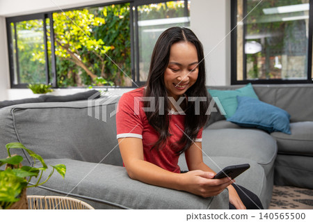 Asian woman sitting on light gray sectional sofa in living room wearing red tee holding smartphone Asian woman sitting on light gray sectional sofa in living room wearing red tee holding smartphone 140565500