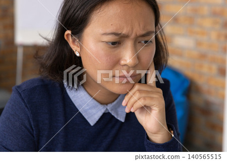 Adult Asian woman touching chin and concentrating at desk in navy sweater and stud earrings Adult Asian woman touching chin and concentrating at desk in navy sweater and stud earrings 140565515