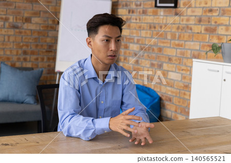 Asian male sitting at wooden table in office, wearing light blue shirt, gesturing near flip chart Asian male sitting at wooden table in office, wearing light blue shirt, gesturing near flip chart 140565521