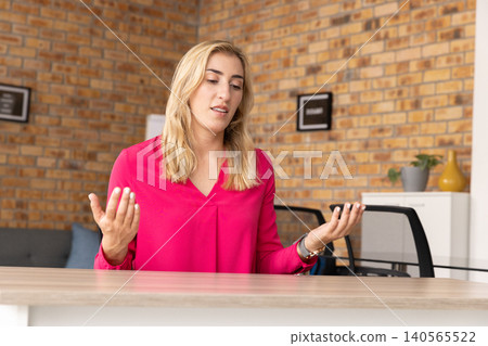 Woman wearing bright pink blouse gesturing while sitting behind wooden table in small office Woman wearing bright pink blouse gesturing while sitting behind wooden table in small office 140565522