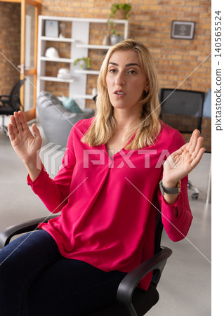 Mid-age woman in magenta blouse sitting in black chair gesturing and talking at office with screen Mid-age woman in magenta blouse sitting in black chair gesturing and talking at office with screen 140565524