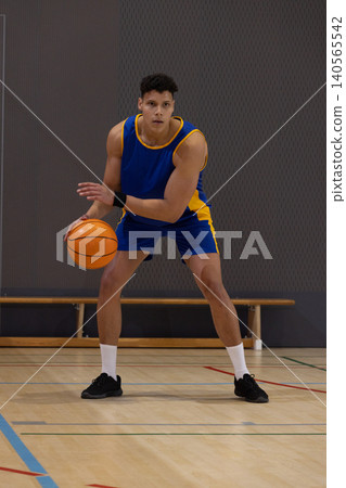 Male 20s in blue jersey holding orange basketball lowering into wide stance on court by bench 140565542