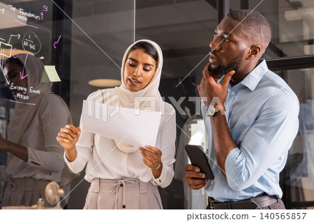 Diverse coworkers standing by glass board at office reading sheet, checking smartphone messages Diverse coworkers standing by glass board at office reading sheet, checking smartphone messages 140565857