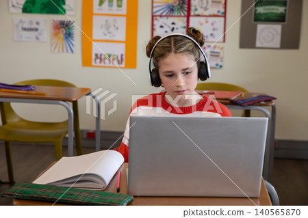 Child girl sitting at school desk studying on laptop with headphones, notebook, pencil case Child girl sitting at school desk studying on laptop with headphones, notebook, pencil case 140565870