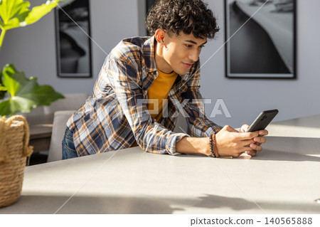 Hispanic man leaning forward on kitchen holding smartphone beside woven basket with plant Hispanic man leaning forward on kitchen holding smartphone beside woven basket with plant 140565888