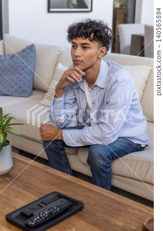 Man in his 20s sitting on sofa at home, remote controls with potted plant nearby Man in his 20s sitting on sofa at home, remote controls with potted plant nearby 140565894