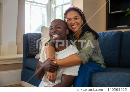 Diverse couple sitting in living room on blue sofa near windows, woman wrapping arms around partner Diverse couple sitting in living room on blue sofa near windows, woman wrapping arms around partner 140565915