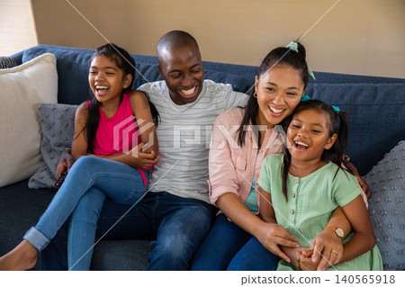 Diverse family of four laughing together on deep blue sofa in living room holding remote control Diverse family of four laughing together on deep blue sofa in living room holding remote control 140565918