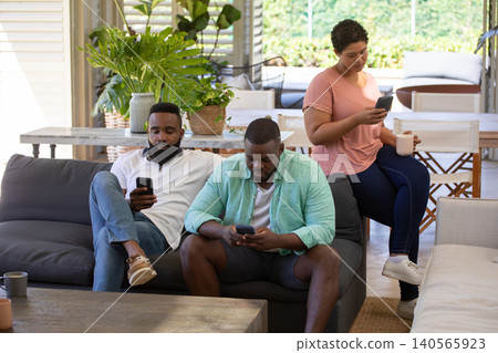 Diverse friends relaxing on grey sofa at home patio scrolling smartphones, holding ceramic mug Diverse friends relaxing on grey sofa at home patio scrolling smartphones, holding ceramic mug 140565923