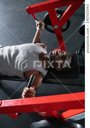 Mid adult African American man lying on bench pushing red chest-press machine weights at gym Mid adult African American man lying on bench pushing red chest-press machine weights at gym 140565943