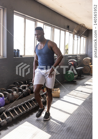African American man standing in gym area wearing sportswear beside kettlebells, plates and fan African American man standing in gym area wearing sportswear beside kettlebells, plates and fan 140565952