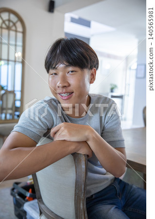 Teenage asian male leaning forward on wooden chair in residential dining room next to arched mirror Teenage asian male leaning forward on wooden chair in residential dining room next to arched mirror 140565981