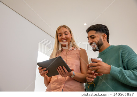 Diverse friends standing at home looking at tablet screen while holding coffee mug Diverse friends standing at home looking at tablet screen while holding coffee mug 140565987
