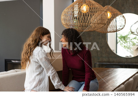 Female partners leaning on wooden dining table at home, smiling across moving box, mirror, lamps Female partners leaning on wooden dining table at home, smiling across moving box, mirror, lamps 140565994