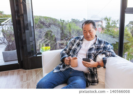 Asian man sipping from ceramic mug, scrolling smartphone on sofa in living room, copy space Asian man sipping from ceramic mug, scrolling smartphone on sofa in living room, copy space 140566008