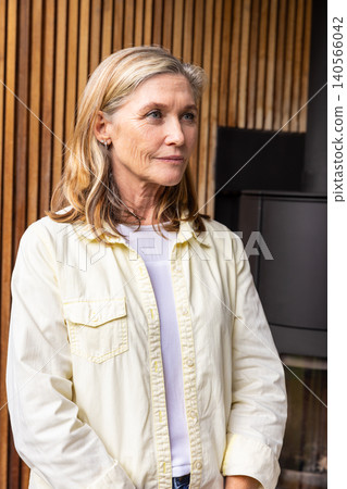 Senior female standing at home by vertical wood slat wall and gazing off next to metal stove Senior female standing at home by vertical wood slat wall and gazing off next to metal stove 140566042