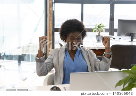 African american woman sitting, raising fists in celebration at office desk with silver laptop African american woman sitting, raising fists in celebration at office desk with silver laptop 140566048