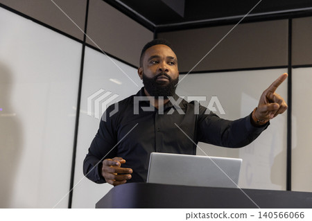 African american man pointing forward while presenting with laptop at podium in meeting room African american man pointing forward while presenting with laptop at podium in meeting room 140566066