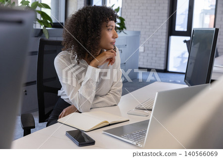 African american woman resting chin on hand and gazing right at white desk in office with notebook African american woman resting chin on hand and gazing right at white desk in office with notebook 140566069