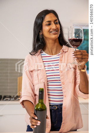 Indian woman holding wine bottle while swirling red wine glass in home kitchen with stove Indian woman holding wine bottle while swirling red wine glass in home kitchen with stove 140566070